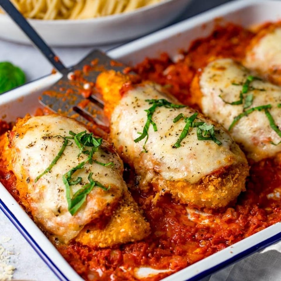 Chicken parmesan with melted mozzarella and fresh basil in a baking dish with a spatula.